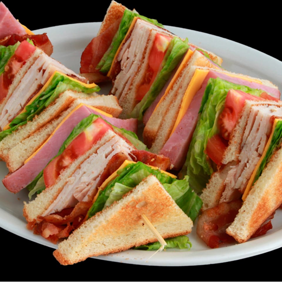A white plate filled with several club sandwiches, cut into triangles, showing layers of meat, cheese, and vegetables.